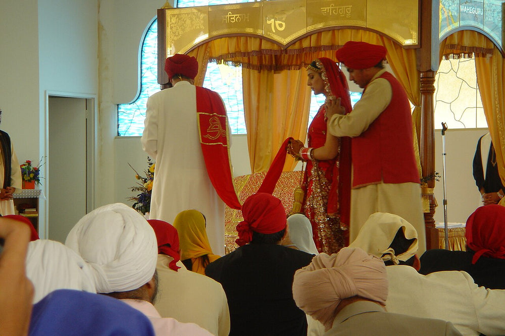 A Sikh bride and groom stand before the Guru Granth Sahib canopy during the Anand Karaj, the palla scarf joining them